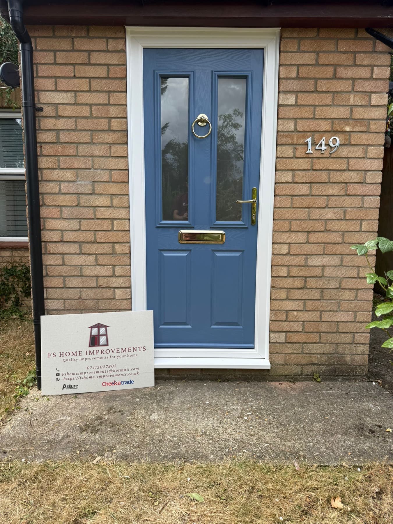 Pigeon blue Hallmark composite front door with gold knocker, letterplate and handle in a white uPVC frame on a buff-brick home