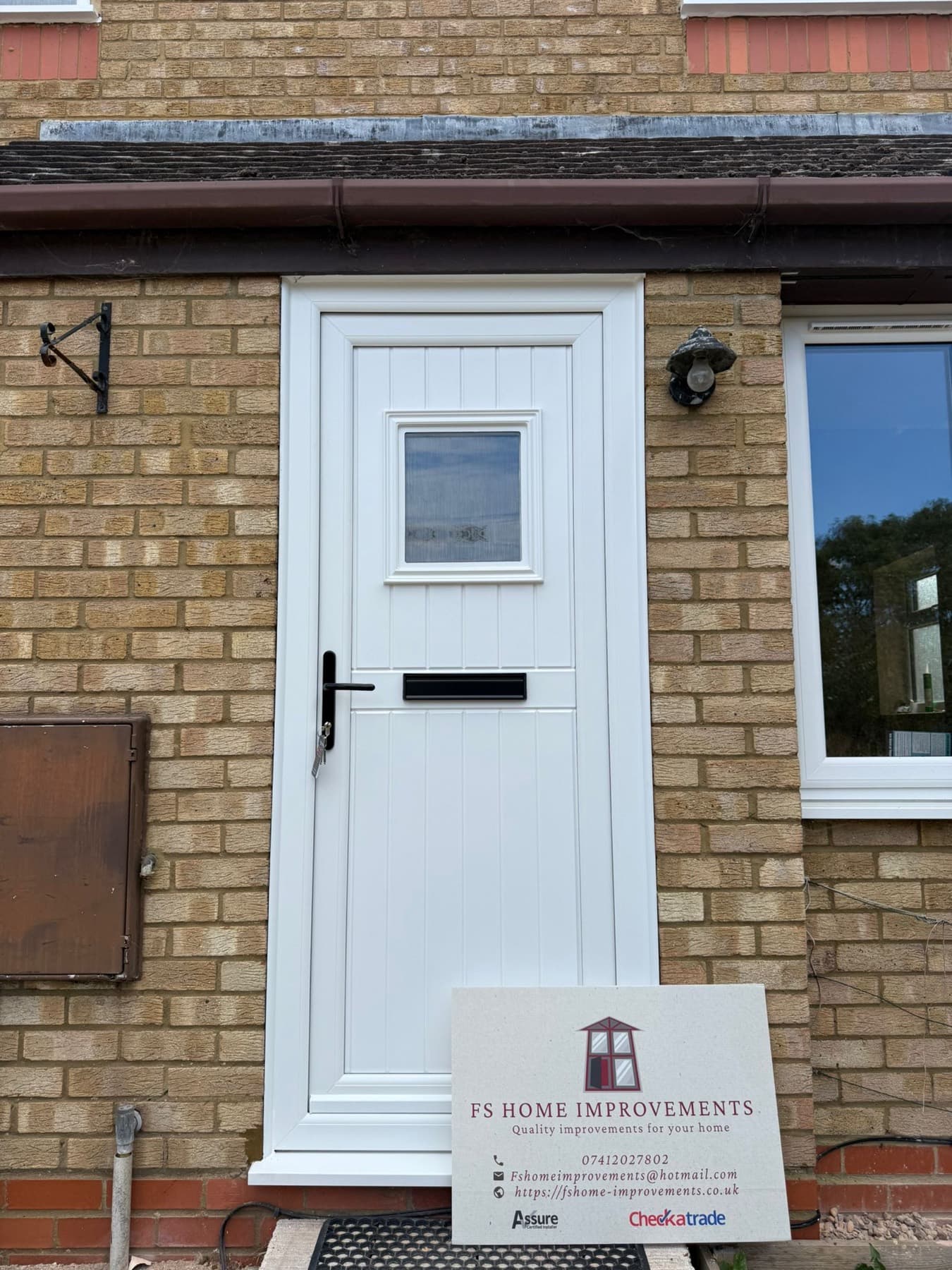 White uPVC stable door with vertical cottage panelling, obscure glazed top light and black handle and letterplate on a buff-brick home